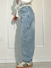 Jeans a vita alta in denim chiaro con dettagli glitterati, tasche laterali, indossati con sneakers bianche e nere, sfondo bianco minimalista.