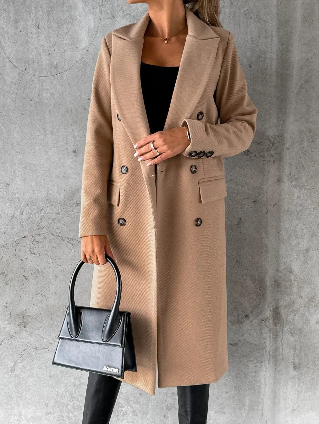 Cappotto lungo beige elegante da donna, doppio petto, abbinato a borsa nera e outfit nero, moda autunnale, sfondo grigio minimalista.