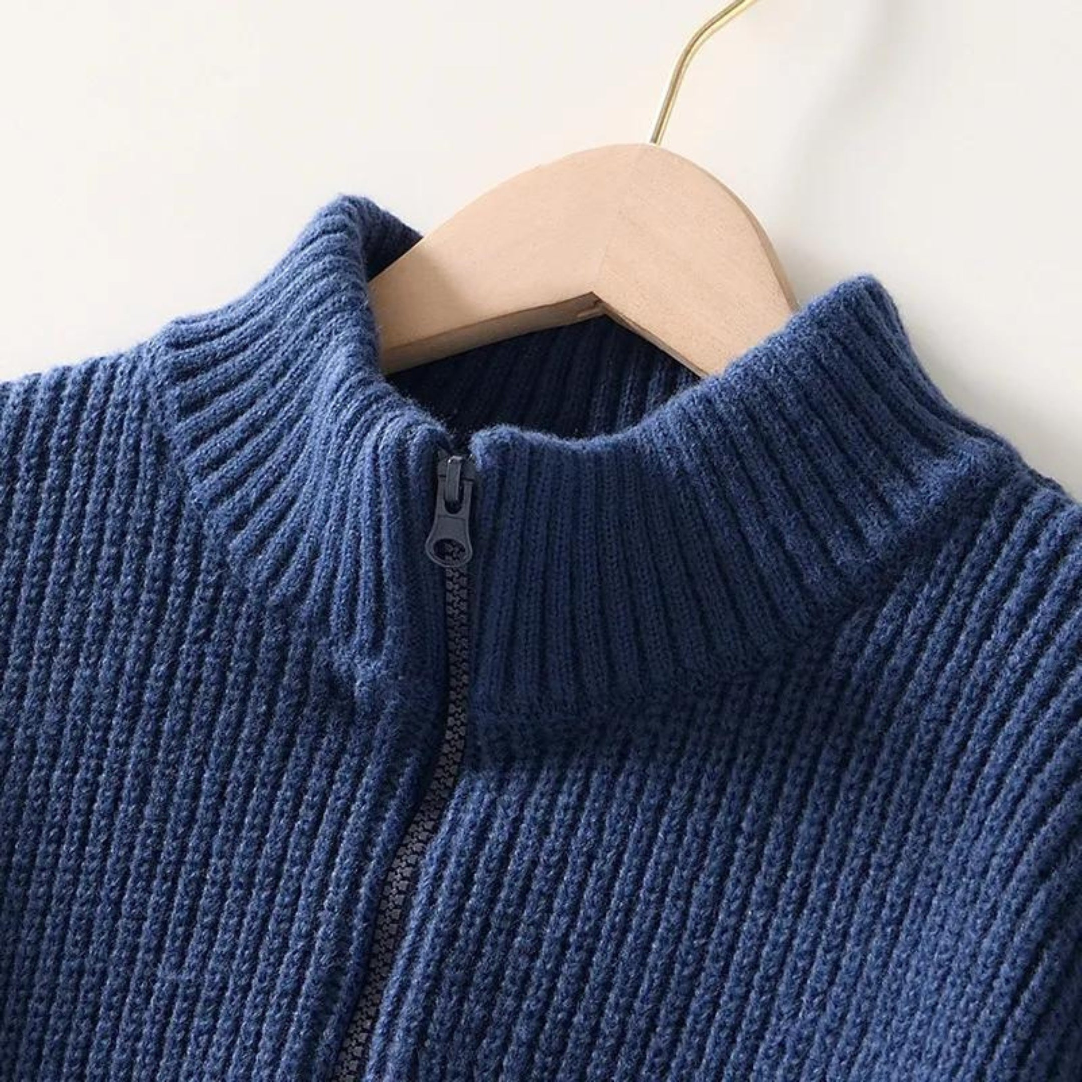 Maglione blu in lana a coste con zip frontale, appeso su gruccia di legno. Design a collo alto, ideale per moda invernale e abbigliamento casual.