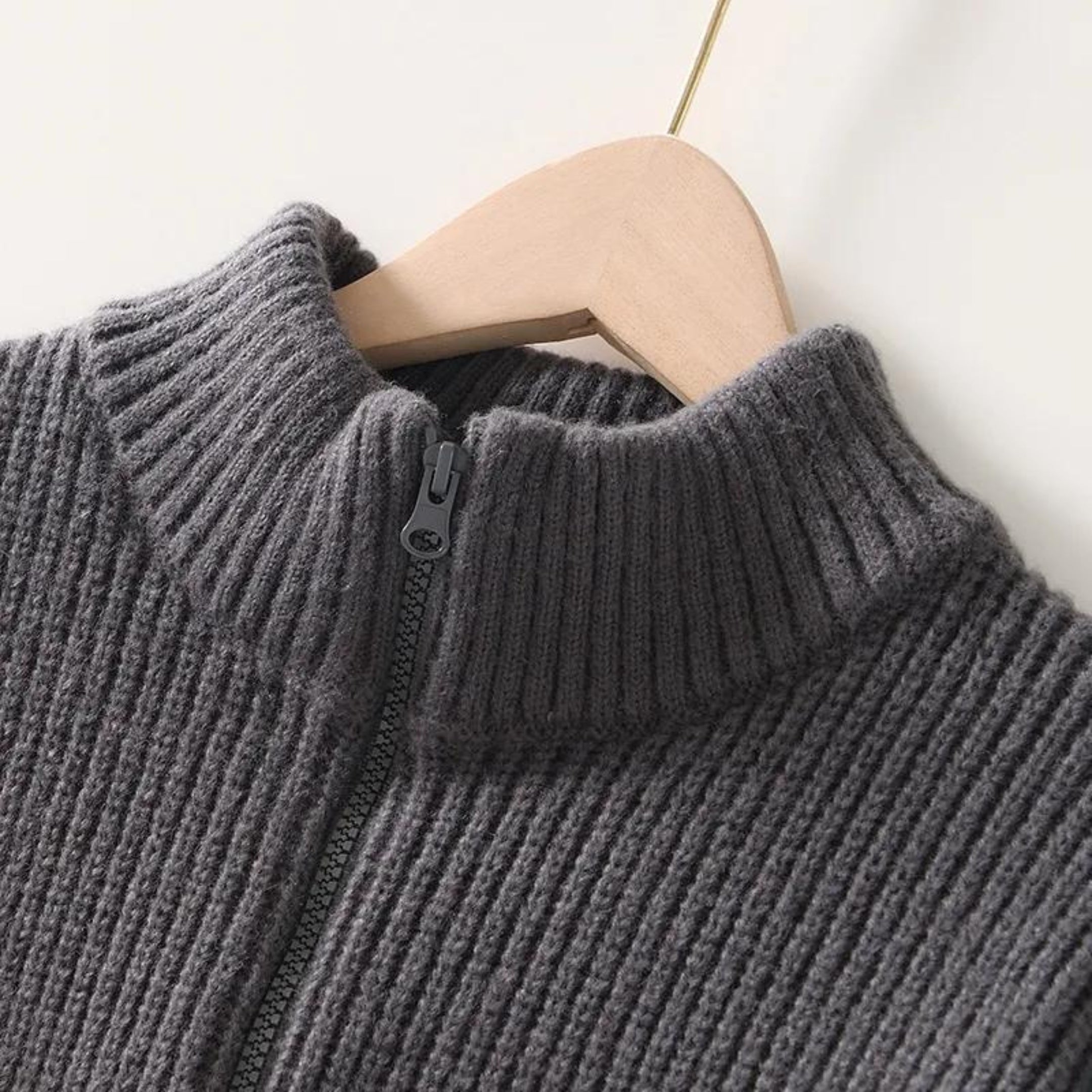 Maglione grigio in lana a coste con zip frontale, collo alto, su appendiabiti in legno. Elegante e caldo, ideale per l'inverno.