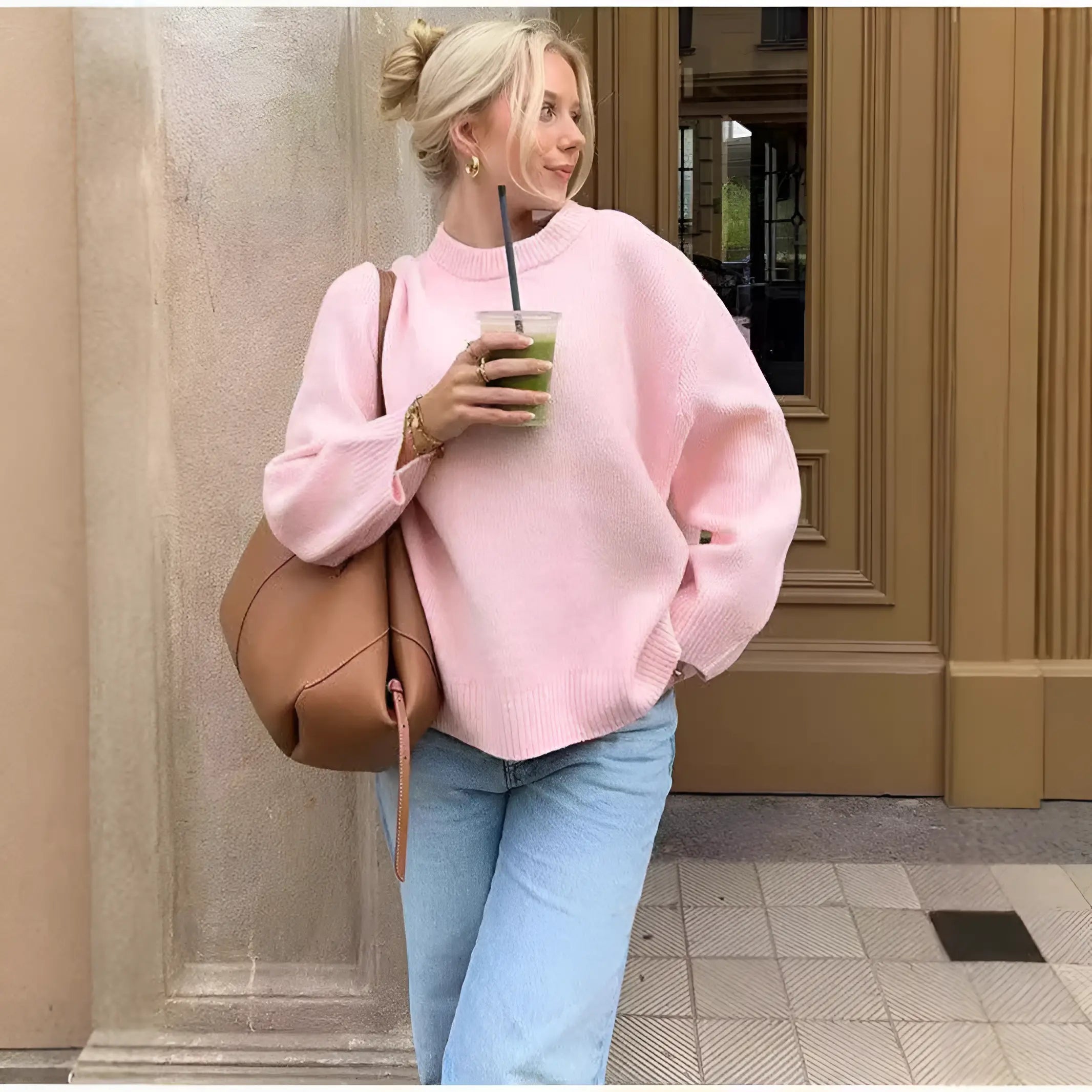 Donna con maglione rosa, jeans chiari e borsa marrone, beve un frullato verde. Moda casual autunnale, look urbano chic.