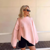 Donna con maglione rosa oversize, occhiali da sole neri e borsa intrecciata, posa all'aperto. Moda autunnale, stile casual chic.