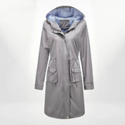 Giacca impermeabile grigia da donna con cappuccio, fodera interna a righe blu, tasche frontali e chiusura a bottoni. Ideale per pioggia e outdoor.