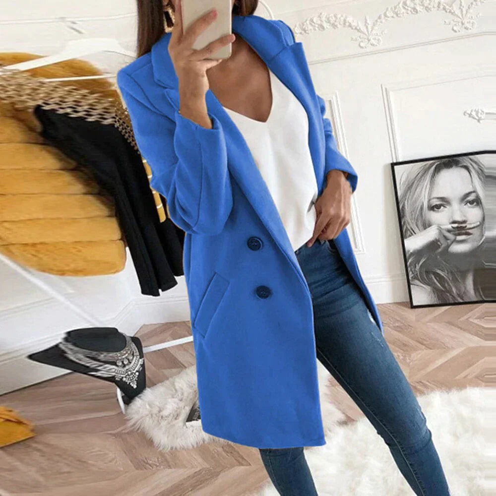 Donna con cappotto blu elegante, jeans aderenti e top bianco, scatta un selfie in una stanza moderna. Moda autunnale, abbigliamento casual chic.