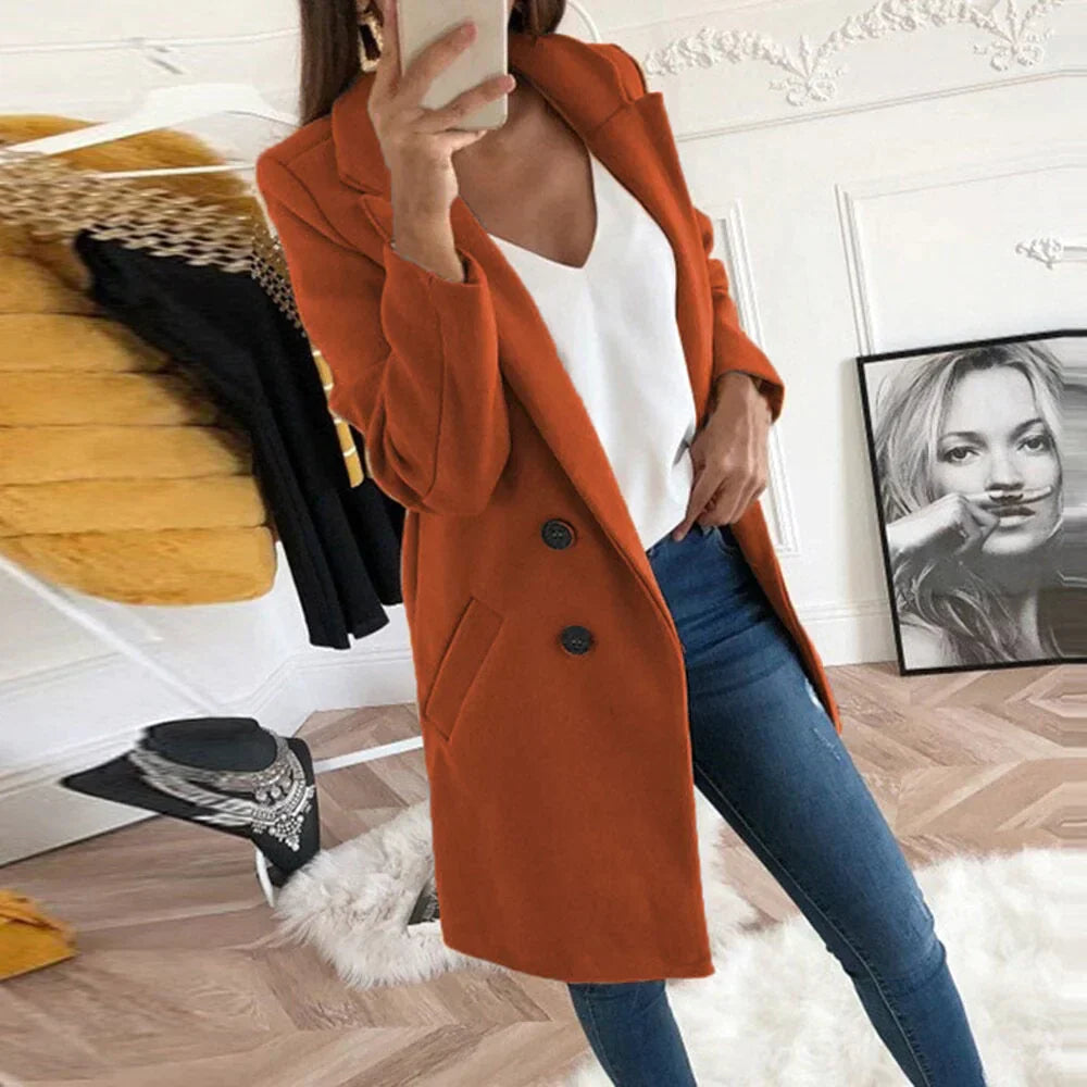 Donna con cappotto arancione elegante, jeans aderenti e top bianco, in una stanza moderna con decorazioni artistiche. Moda autunnale chic.