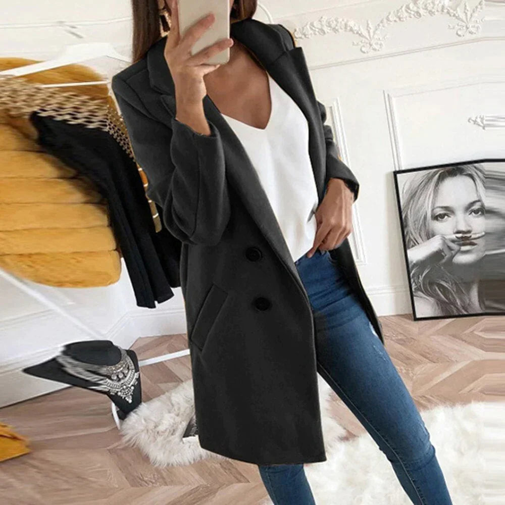 Donna in cappotto nero elegante, jeans aderenti e top bianco, scatta selfie in salotto moderno. Moda autunnale, abbigliamento casual chic.