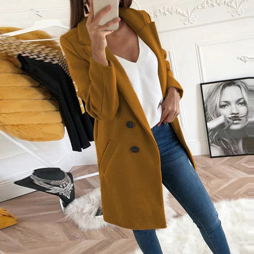Donna con cappotto lungo color senape, jeans aderenti e top bianco, in una stanza moderna con decorazioni eleganti e specchio. Moda autunnale chic.