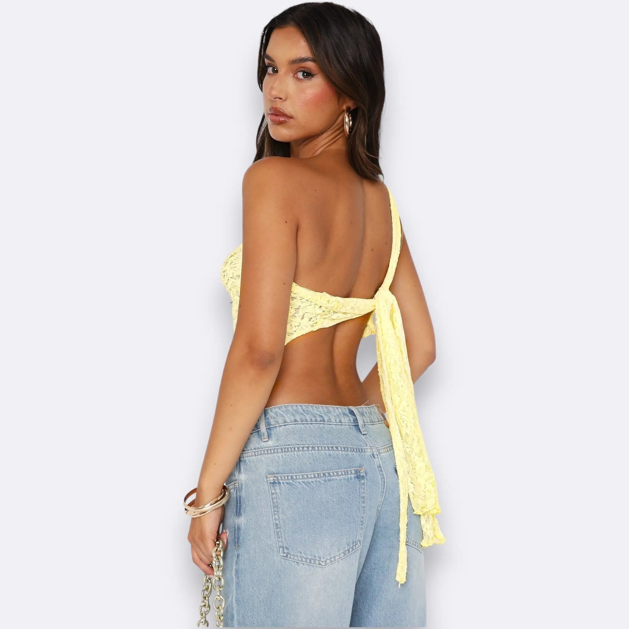 Donna con top giallo monospalla in pizzo, schiena scoperta, jeans a vita alta, orecchini a cerchio, sfondo bianco. Moda estiva, abbigliamento casual.