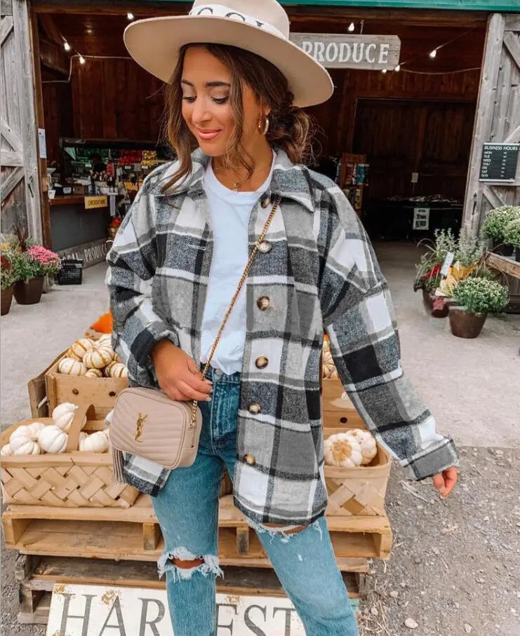 Donna con cappotto a quadri grigio e bianco, jeans strappati, borsa a tracolla beige, cappello beige, davanti a un mercato agricolo con zucche.
