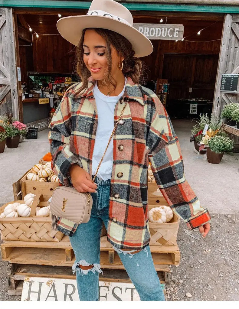 Donna con cappotto a quadri, jeans strappati e cappello beige davanti a un mercato agricolo. Borsa a tracolla beige, zucche e fiori sullo sfondo.
