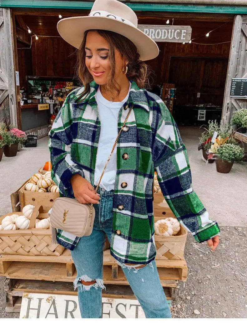 Donna con cappello beige e camicia a quadri verde e blu, davanti a un mercato agricolo con zucche. Moda autunnale, stile casual chic.