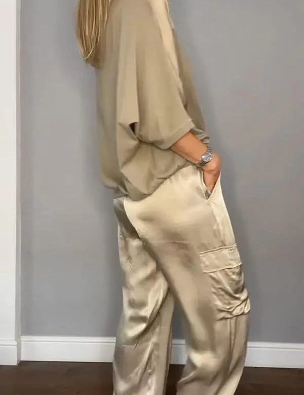 Donna in abbigliamento elegante beige, indossa pantaloni di seta e maglia morbida, posa sorridente su sfondo grigio. Moda femminile chic.