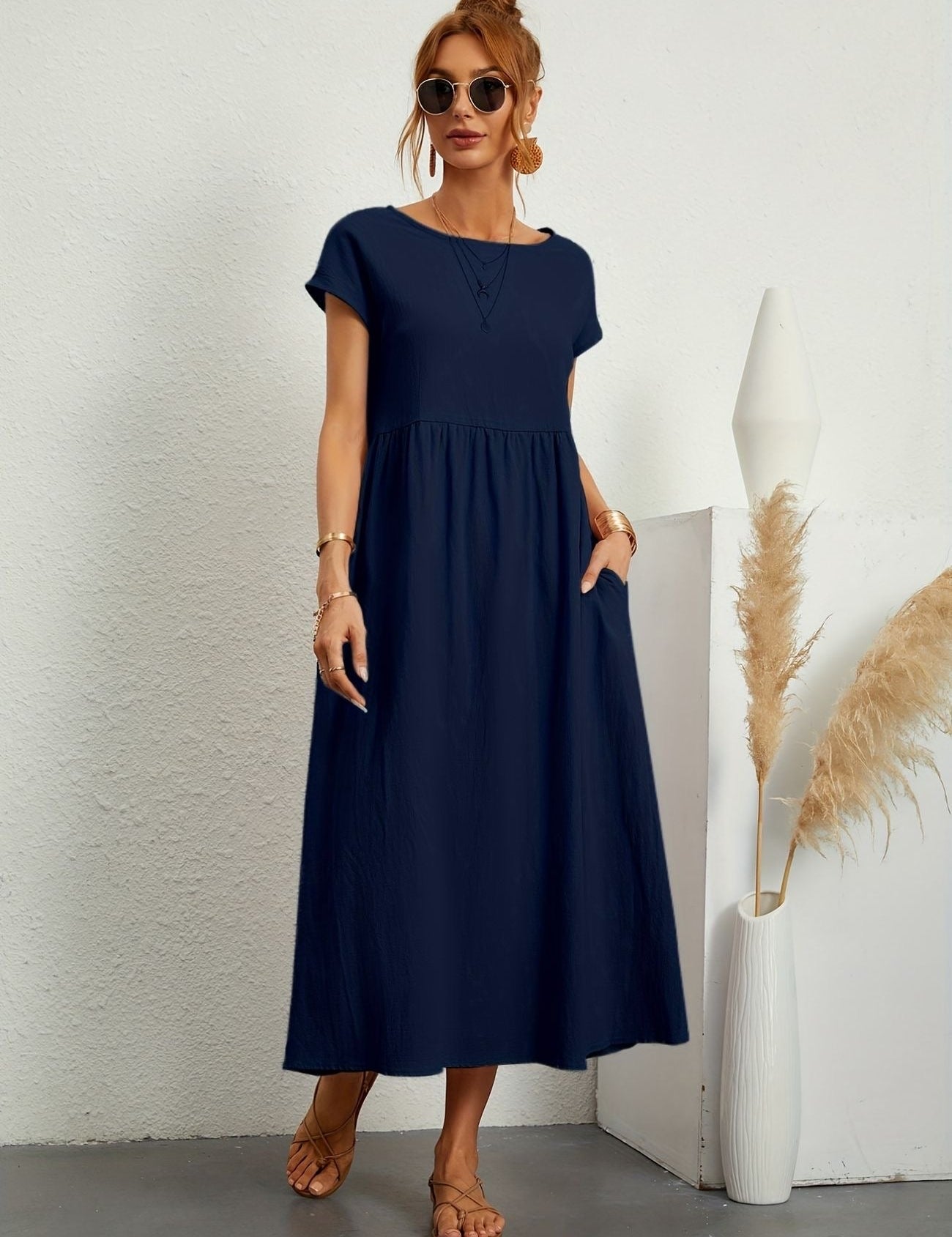 Donna con abito lungo blu scuro, occhiali da sole, sandali beige, accanto a vaso bianco con pampas. Moda estiva elegante e casual.