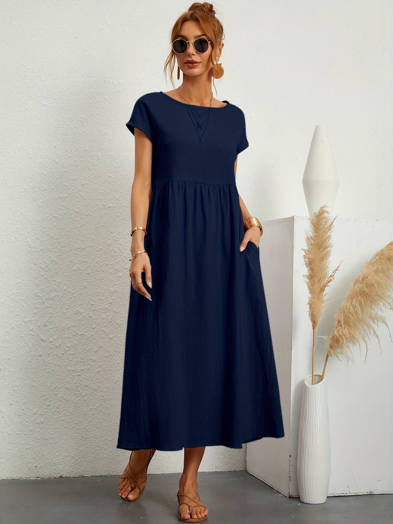 Donna con abito lungo blu scuro, occhiali da sole, sandali beige, accanto a vaso bianco con pampas. Moda estiva elegante e casual.