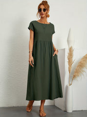 Donna con abito lungo verde oliva, occhiali da sole, sandali beige, accanto a vaso bianco con pampas. Moda estiva casual elegante.