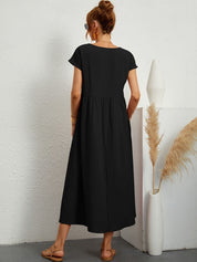 Donna con abito lungo nero, sandali beige, capelli raccolti, occhiali da sole, accanto a vaso bianco con pampas. Moda estiva elegante e casual.