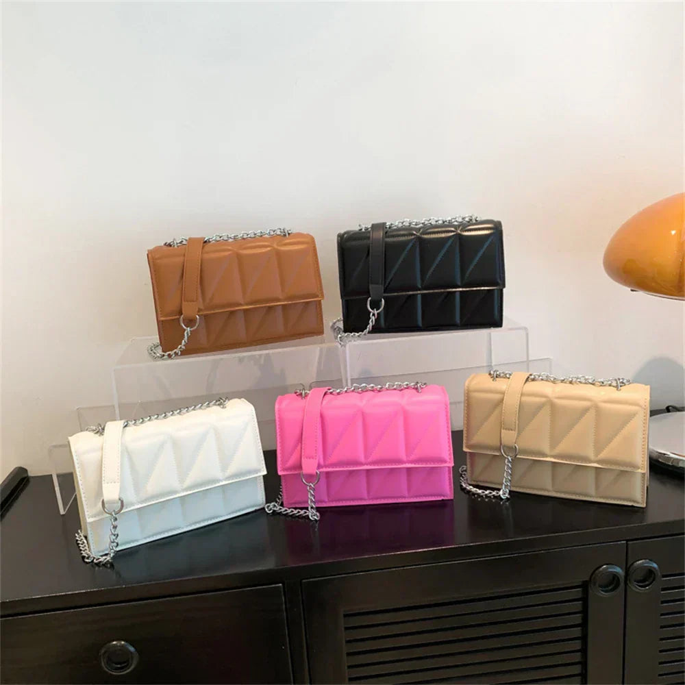 Cinque borse a tracolla trapuntate in pelle PU, colori assortiti: marrone, nero, bianco, rosa, beige, con catena argentata, esposte su scaffale.