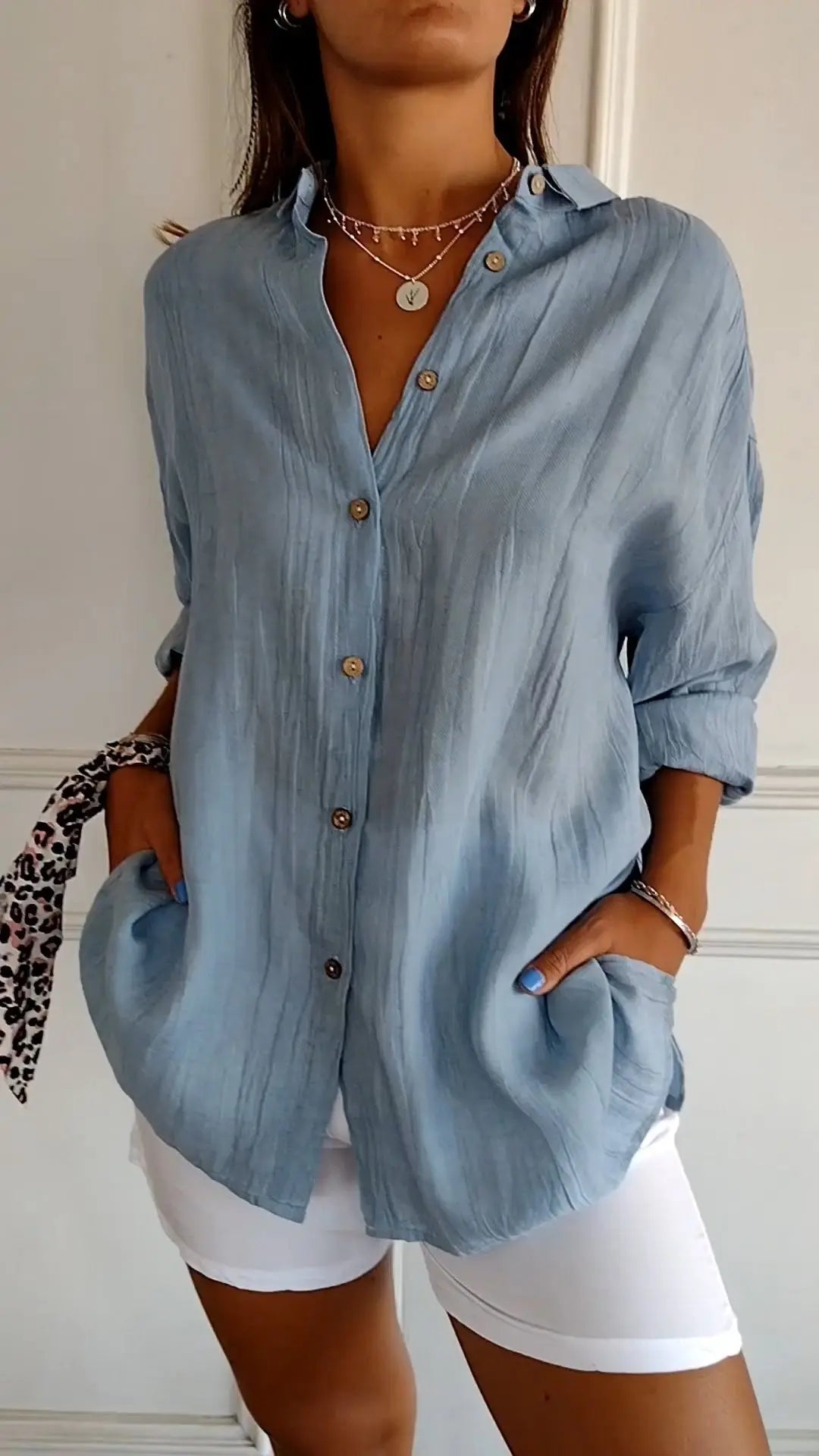 Camicia donna azzurra in lino con bottoni, maniche lunghe arrotolate, abbinata a pantaloni bianchi. Stile casual elegante, moda estiva.