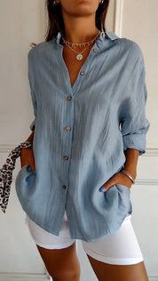 Camicia donna azzurra in lino con bottoni, maniche lunghe arrotolate, abbinata a pantaloni bianchi. Stile casual elegante, moda estiva.
