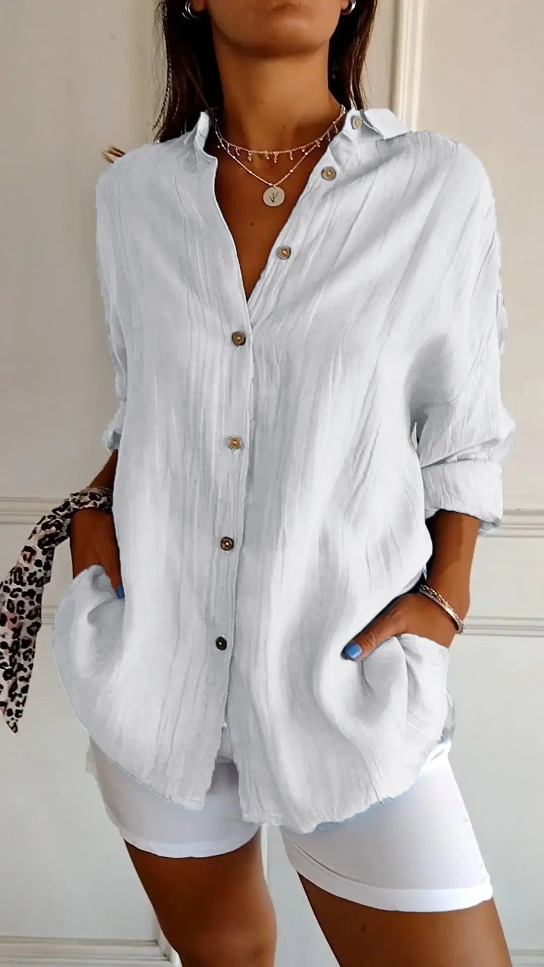 Camicia bianca da donna in lino, maniche lunghe, bottoni marroni, tasche frontali, stile casual elegante, abbinata a pantaloni bianchi.