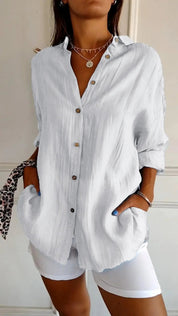 Camicia bianca da donna in lino, maniche lunghe, bottoni marroni, tasche frontali, stile casual elegante, abbinata a pantaloni bianchi.