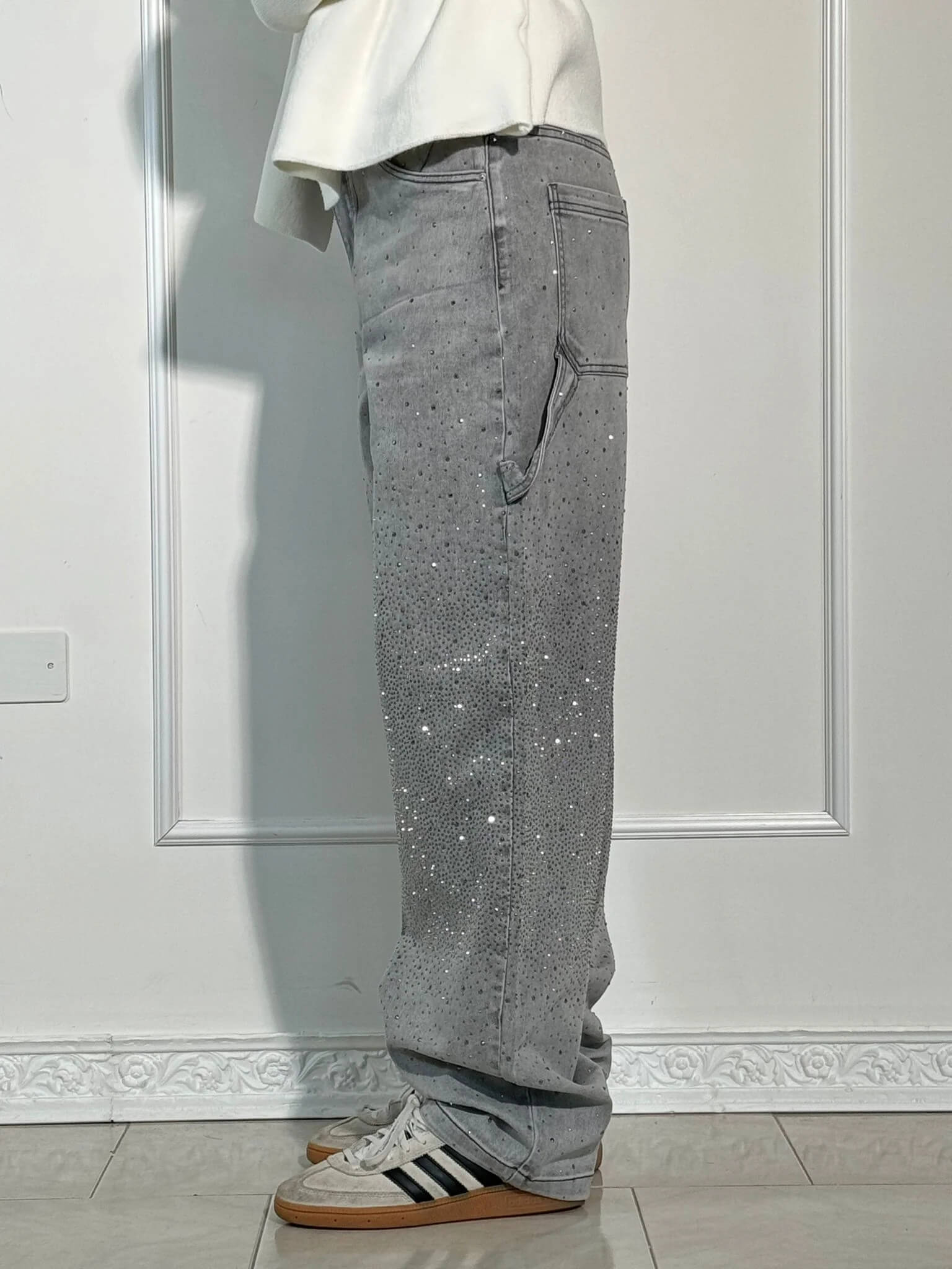 Pantaloni grigi in denim con strass, stile casual, indossati con scarpe sportive Adidas. Design moderno, ideale per moda urbana e streetwear.