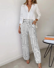 Donna con camicia bianca e pantaloni a palazzo grigi con motivo geometrico, in piedi accanto a un tavolo moderno con libri. Moda elegante e contemporanea.