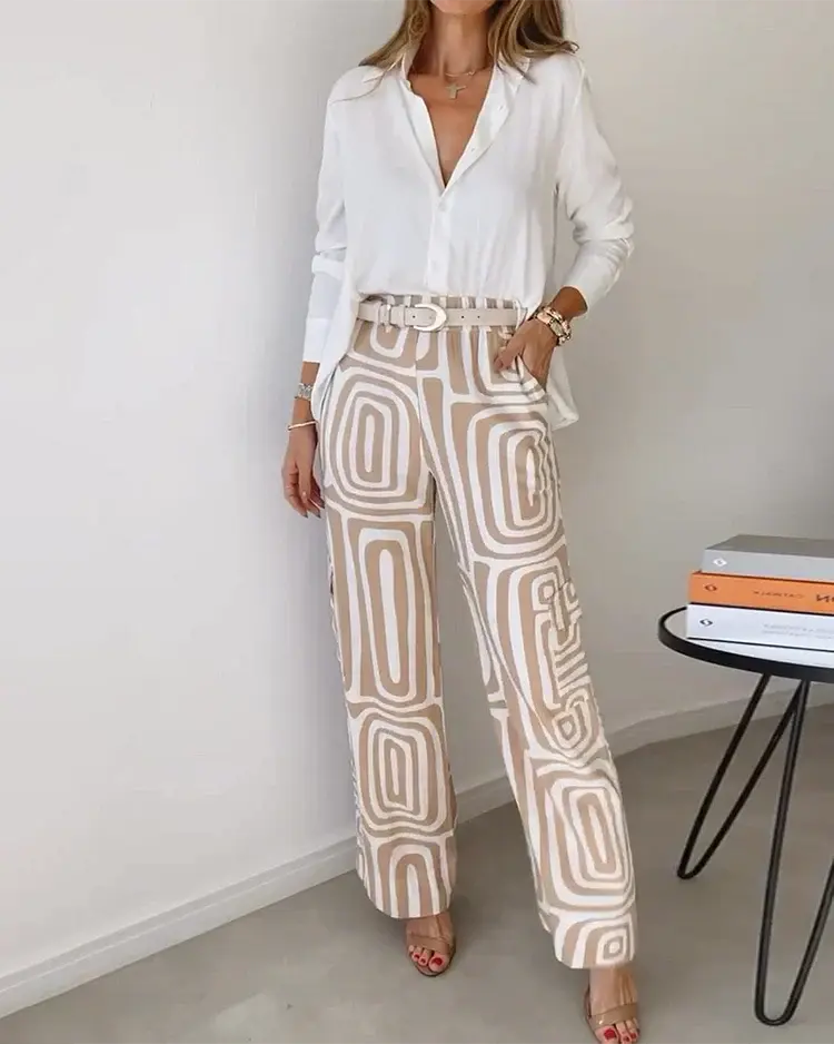 Donna in camicia bianca e pantaloni a fantasia geometrica beige, in piedi accanto a un tavolino con libri. Look elegante e moderno per l'ufficio.