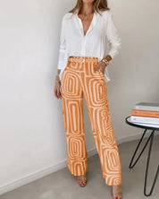 Donna in camicia bianca e pantaloni arancioni a motivi geometrici, in piedi accanto a un tavolo con libri. Moda casual elegante, abbigliamento femminile.