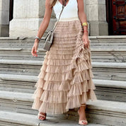 Donna con gonna lunga a balze beige, top bianco senza maniche, borsa a tracolla grigia, sandali eleganti, su gradini di pietra. Moda estiva chic.