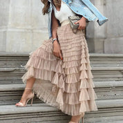 Donna con gonna lunga a balze beige, top bianco, giacca di jeans e sandali con tacco. Moda elegante, outfit primaverile, accessori chic.