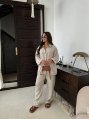 Donna con occhiali da sole indossa completo beige in lino, borsa a tracolla marrone, sandali, in soggiorno moderno con mobili in legno scuro.