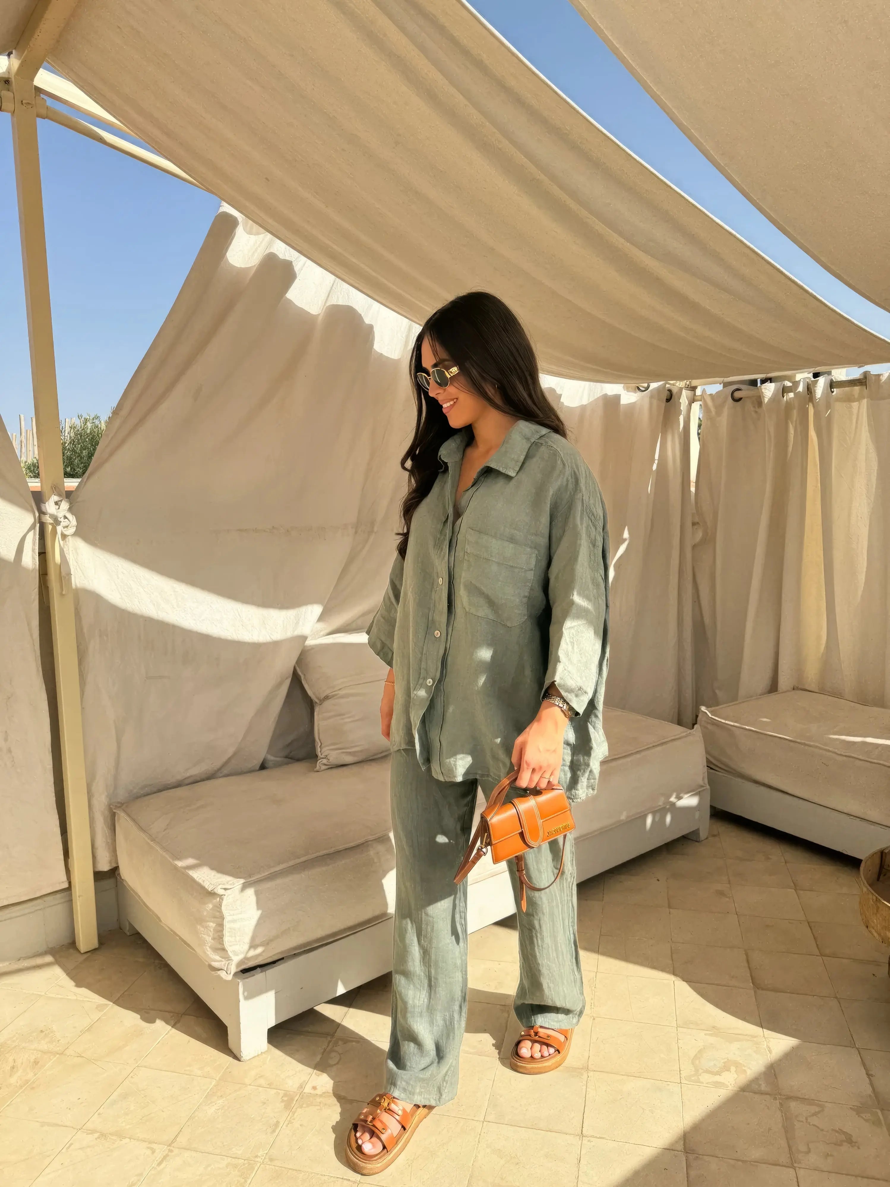Donna con completo verde in lino, occhiali da sole e borsa arancione, in terrazza soleggiata. Moda estiva, abbigliamento casual elegante.