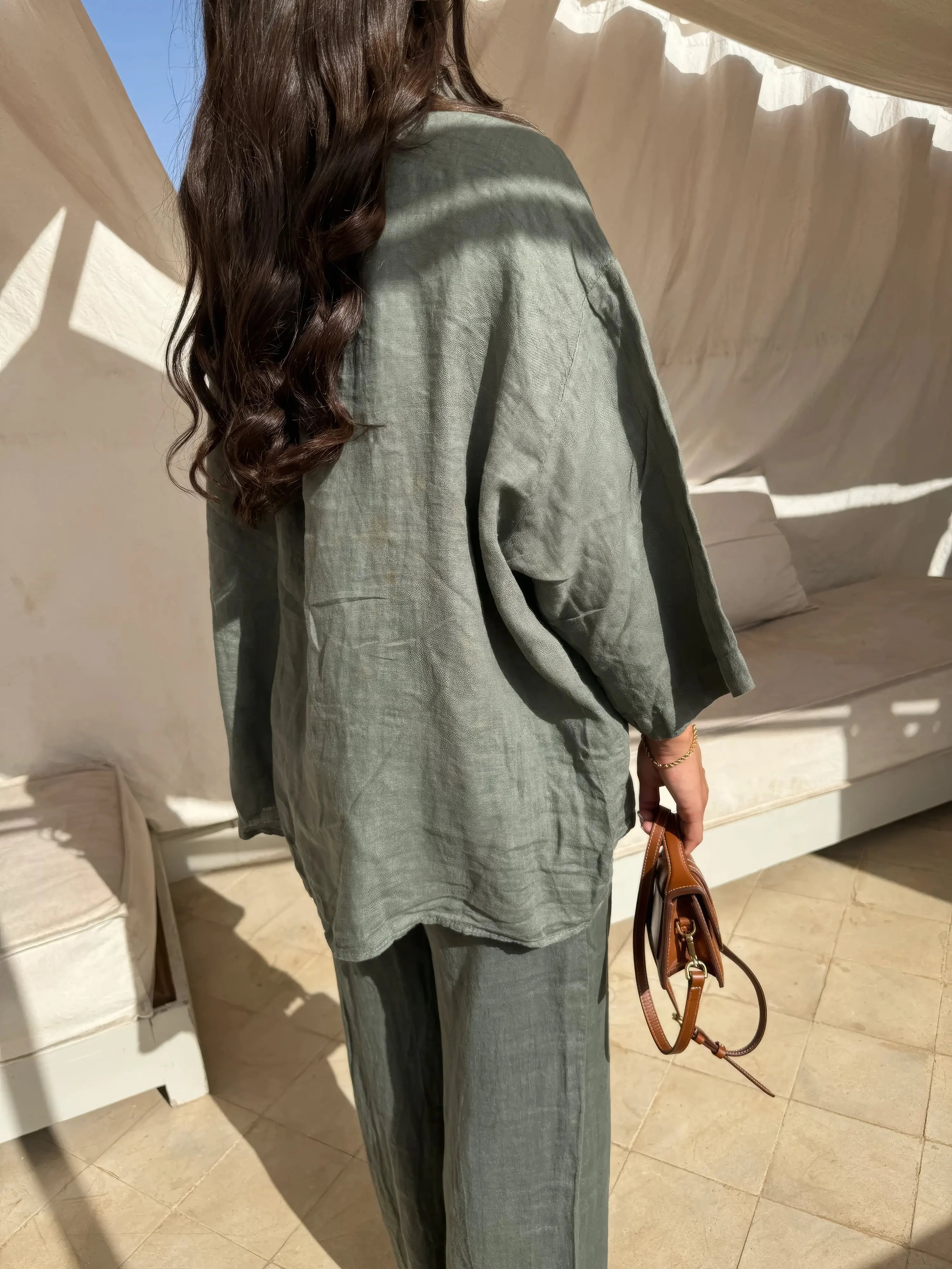 Donna con abito in lino verde oliva, capelli lunghi ondulati, tiene una borsa marrone. Moda estiva, abbigliamento casual elegante.