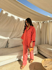 Donna con camicia e pantaloni rossi, occhiali da sole, borsa a tracolla marrone, sandali, in terrazza con tende bianche. Moda estiva casual.