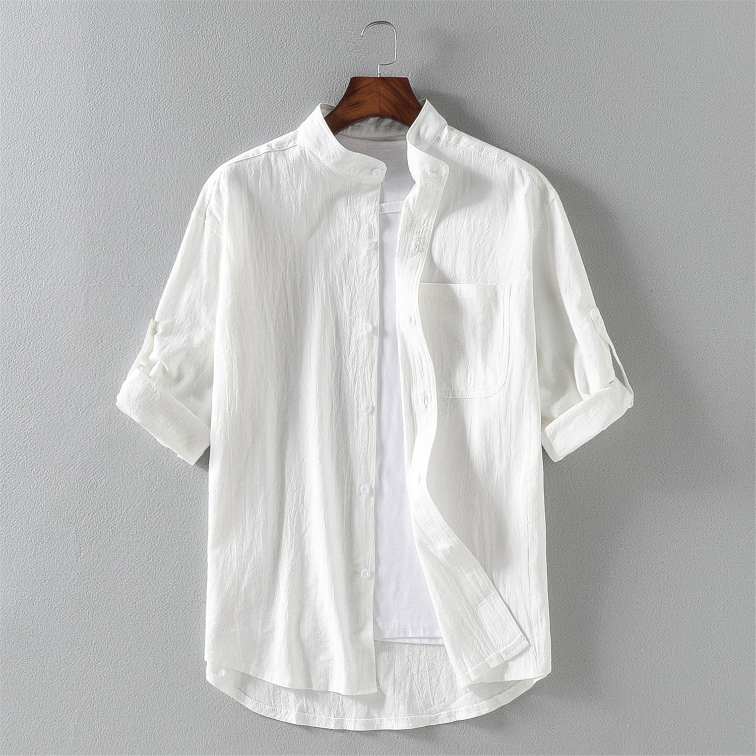Camicia bianca da uomo in cotone, maniche lunghe arrotolate, colletto alla coreana, design casual elegante, appesa su gruccia di legno.