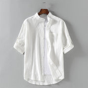Camicia bianca da uomo in cotone, maniche lunghe arrotolate, colletto alla coreana, design casual elegante, appesa su gruccia di legno.