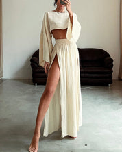 Abito lungo beige con spacco alto e maniche ampie, indossato da una donna in un interno minimalista. Moda elegante, abbigliamento estivo.