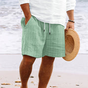 Uomo in spiaggia con pantaloncini verdi a righe, camicia bianca e cappello di paglia. Moda estiva, abbigliamento casual mare.