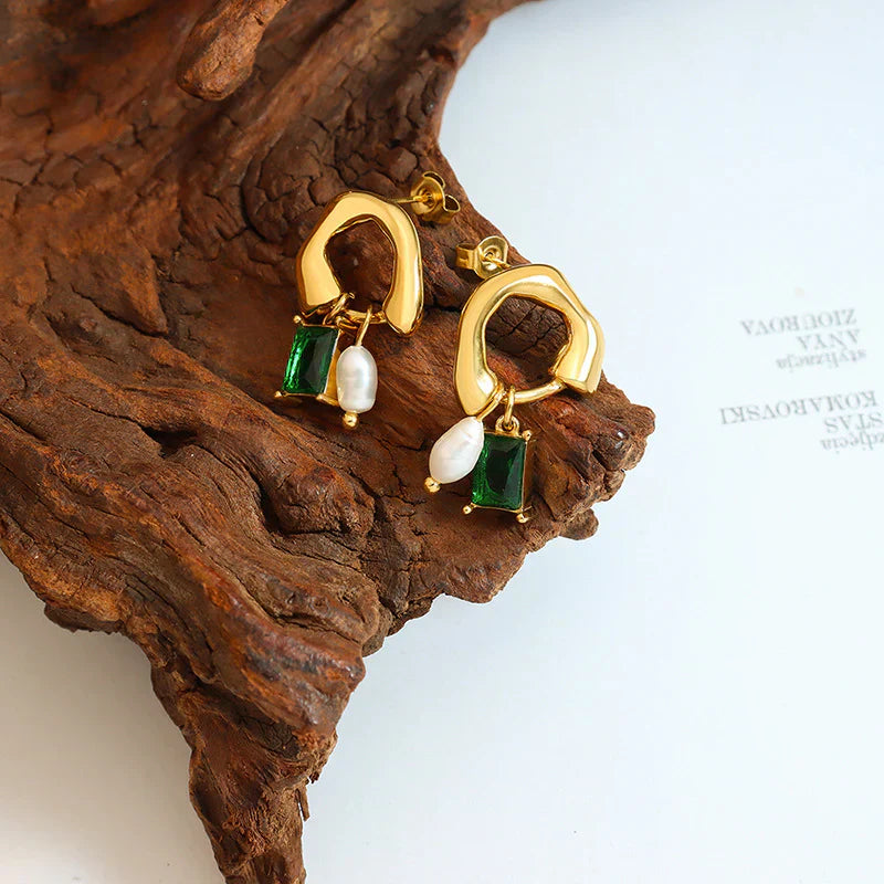Orecchini pendenti in oro con perle bianche e pietre verdi su sfondo di legno. Gioielli eleganti, accessori moda donna.