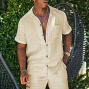 Uomo in camicia beige di lino, maniche corte, con pantaloni coordinati, indossa collana e orologio dorato, sfondo verde naturale. Moda estiva casual.
