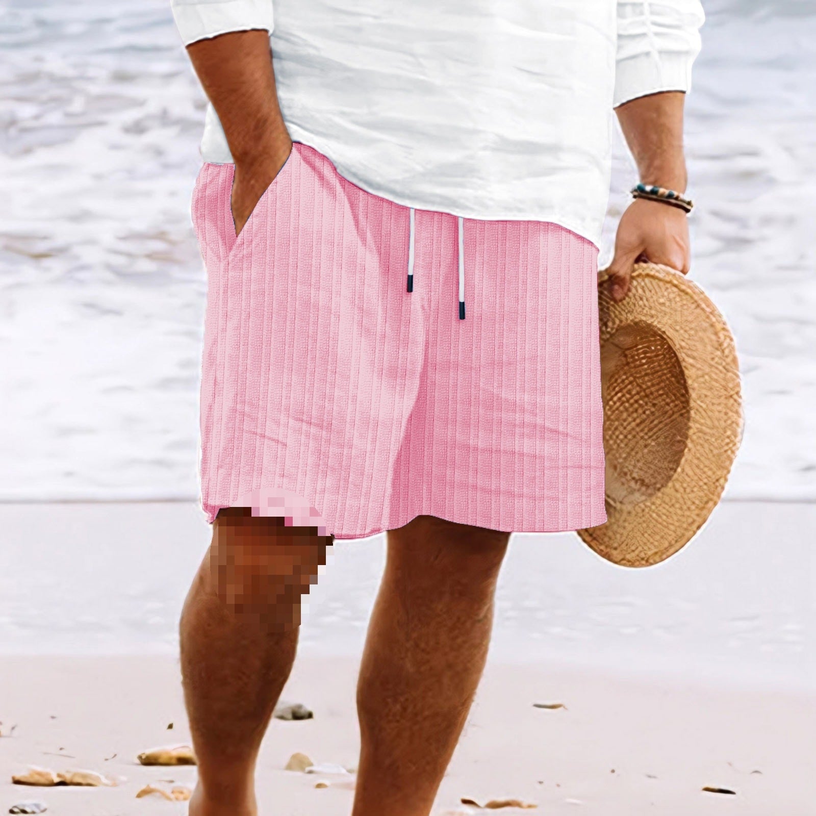 Shorts da spiaggia rosa a righe, uomo in camicia bianca, cappello di paglia in mano, sabbia e mare sullo sfondo. Moda estiva, abbigliamento casual.