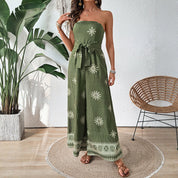 Donna in tuta verde con motivi floreali bianchi, sandali marroni, accanto a pianta tropicale e sedia in vimini. Moda estiva elegante e casual.