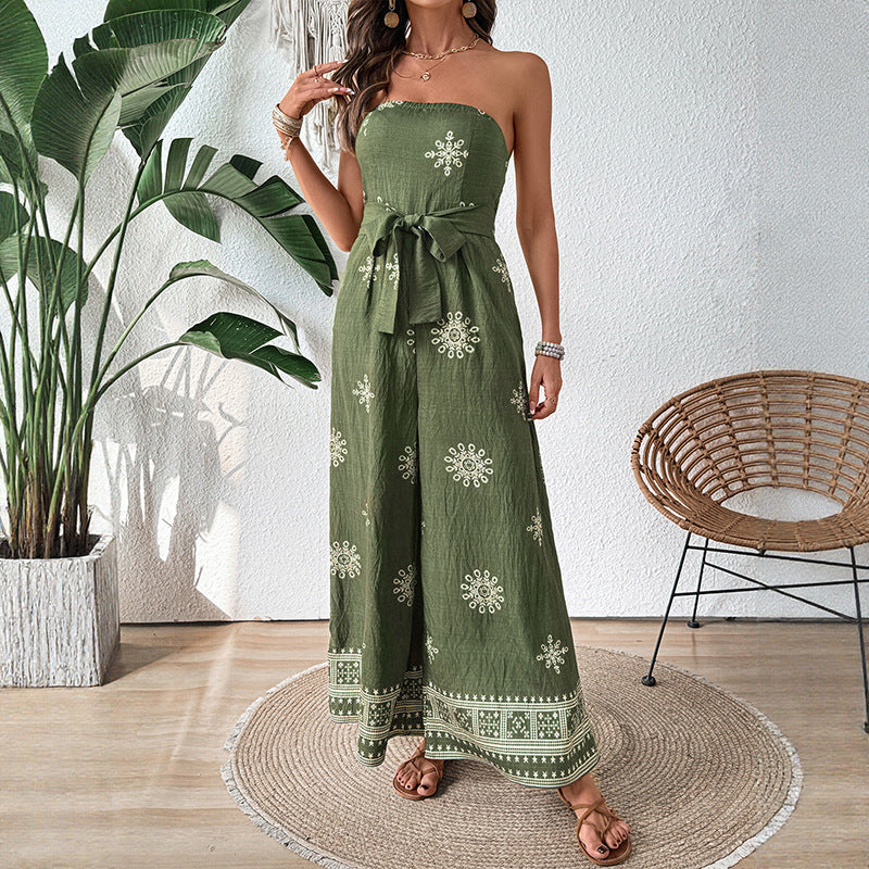 Donna in tuta verde con motivi floreali bianchi, sandali marroni, accanto a pianta tropicale e sedia in vimini. Moda estiva elegante e casual.