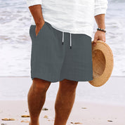 Uomo in spiaggia con pantaloncini grigi e camicia bianca, tiene un cappello di paglia. Moda estiva, abbigliamento casual mare.