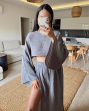 Donna in abito coordinato grigio con top a maniche larghe e gonna lunga con spacco, in soggiorno moderno. Moda femminile, abbigliamento estivo elegante.