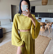 Donna con abito coordinato giallo senape, top a maniche lunghe e gonna plissettata, in soggiorno moderno. Moda estiva, abbigliamento casual elegante.