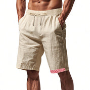 Pantaloncini uomo beige in lino, elastico in vita, tasche laterali, dettaglio rosso geometrico sull'orlo. Abbigliamento estivo casual.