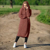 Donna con cappotto lungo a maglia marrone, cappuccio, in piedi su strada sterrata. Moda autunnale, abbigliamento casual, stile confortevole.