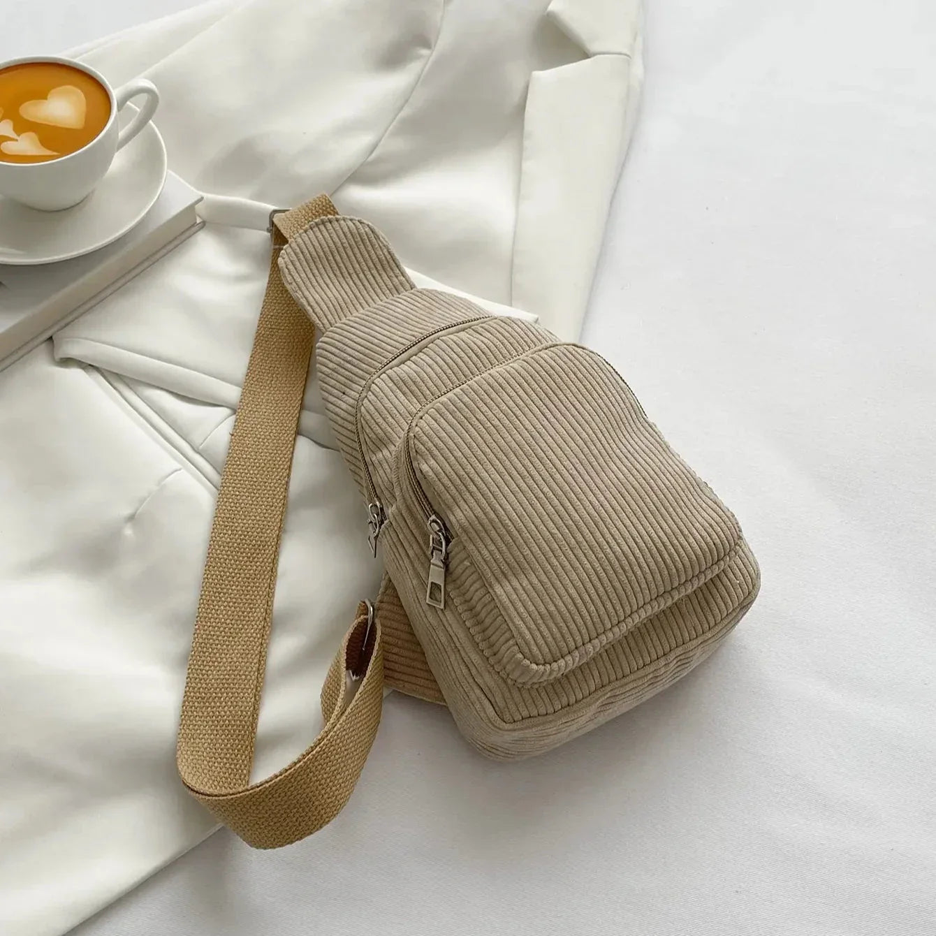 Zaino a tracolla beige in tessuto a coste su sfondo bianco, accanto a una tazza di cappuccino. Accessorio moda casual, ideale per ogni giorno.